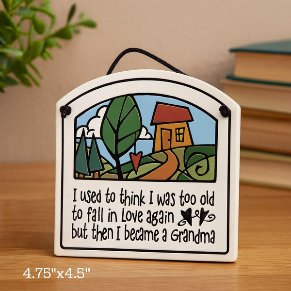 Ceramic wall tile with cottage illustration and the quote ‘I used to think I was too old to fall in love again but then I became a Grandma,’ displayed on a wooden surface beside books.