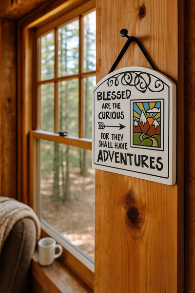 Ceramic wall tile reading ‘Blessed are the curious for they shall have adventures’ hanging inside a cozy wood cabin with forest view.