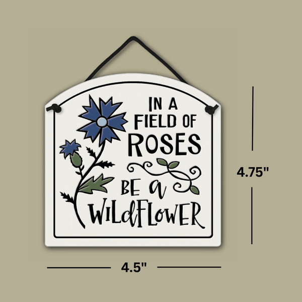Ceramic wall tile with blue wildflower design and the quote ‘In a field of roses be a wildflower,’ shown with dimensions 4.75 inches tall and 4.5 inches wide.