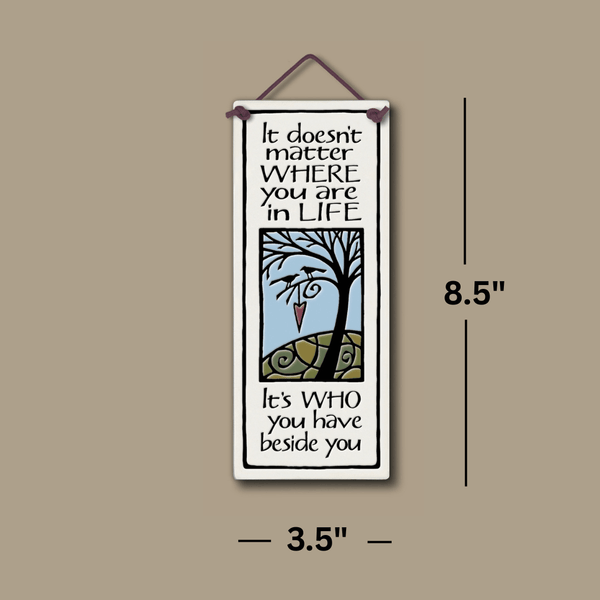 Spooner Creek ceramic tall tile with illustrated tree, heart accent, and the phrase It doesn’t matter where you are in life, it’s who you have beside you, shown with hanging cord and product dimensions 3.5 x 8.5 inches.