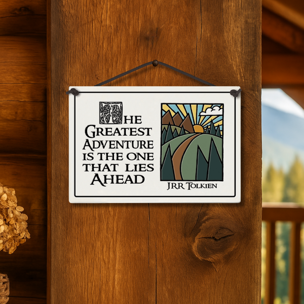 Spooner Creek ceramic tile hanging on a rustic cabin wood post with quote “The greatest adventure is the one that lies ahead – JRR Tolkien,” mountain sunrise artwork, cozy mountain cabin setting.