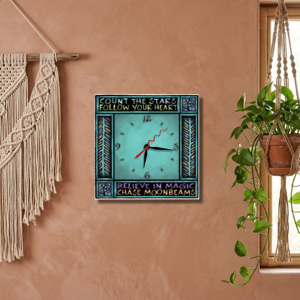 Believe in Magic teal square wood wall clock hanging on a soft blush wall next to a hanging plant. 