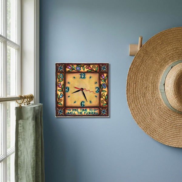 Floral square wood wall clock hanging on a dusty blue wall beside a straw garden hat and a sunny window