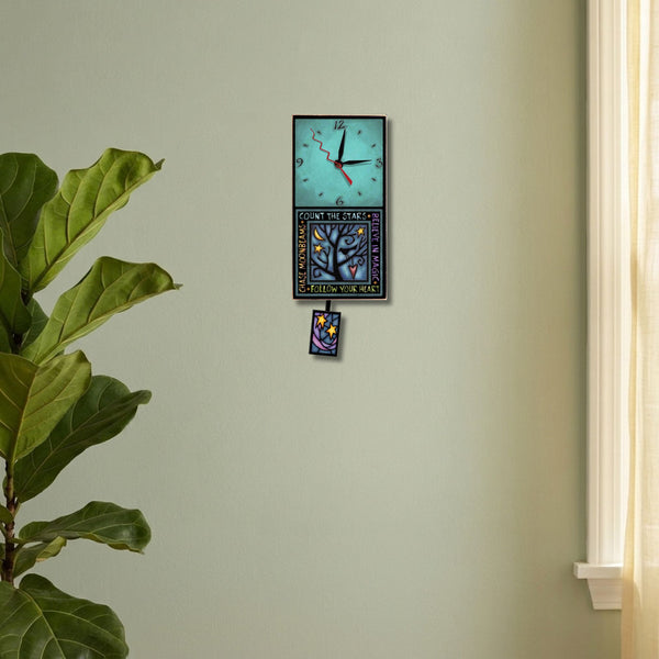 Follow Your Heart handmade pendulum wall clock hanging on a sage green next to a plant and a window.