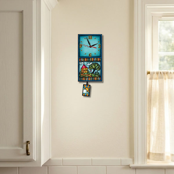 House and tree handmade pendulum wall clock hanging on a kitchen wall between a white cabinet and a sunny window