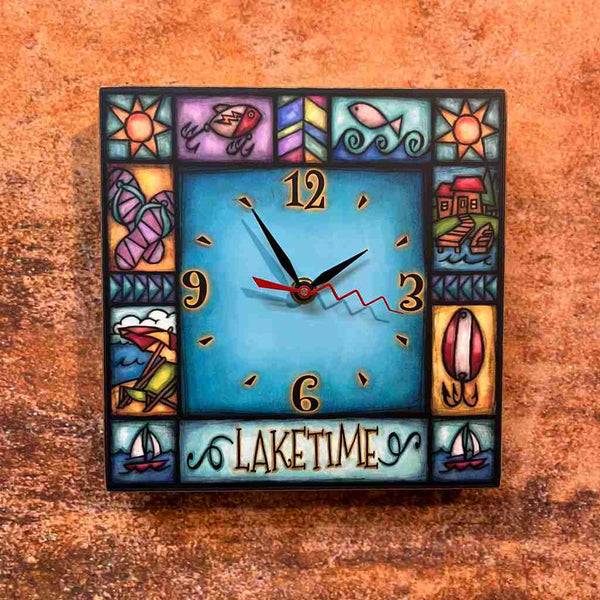 Close-up of the Laketime lake house square wood wall clock on a textured wall, showing the turquoise face, fishing lures, sailboats, and lake cabin border detail