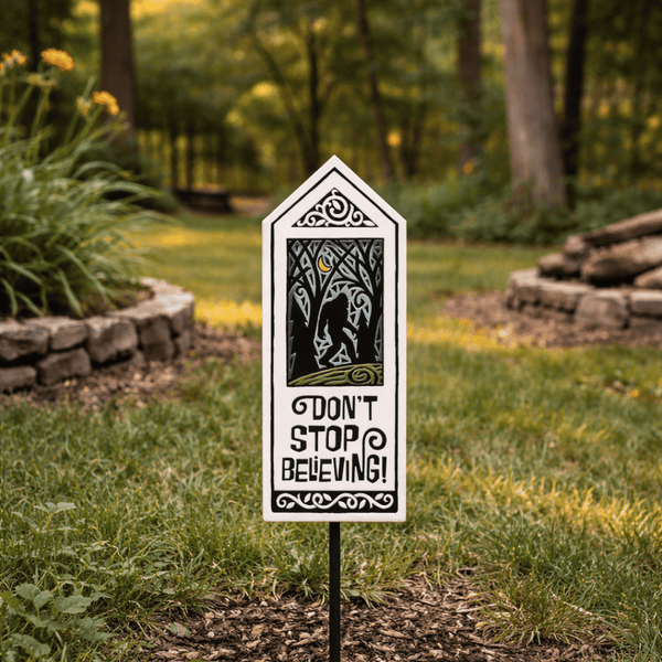 Sasquatch ceramic garden tile stake displayed outdoors in a grassy yard with wooded forest background, hand glazed Bigfoot garden decor with Dont Stop Believing quote