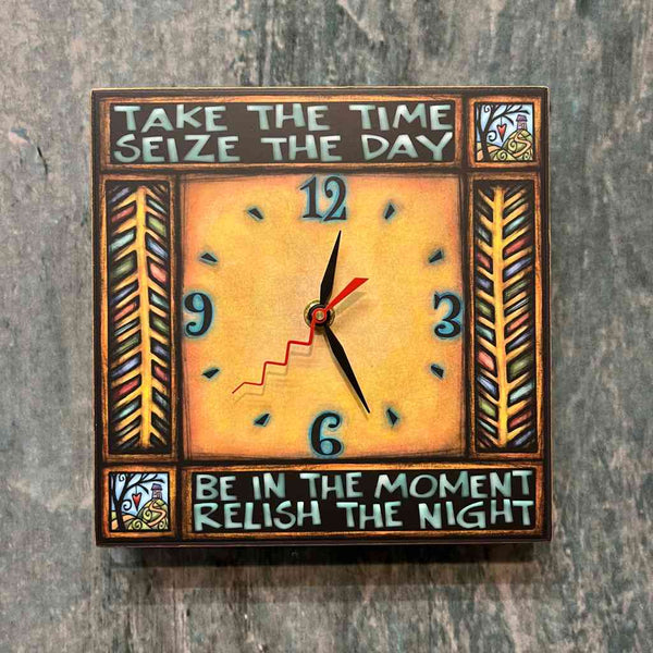 Close-up of the Take the Time seize the day square wood wall clock on a textured wall, showing the golden face, hand-lettered quote, and feather panel border detail