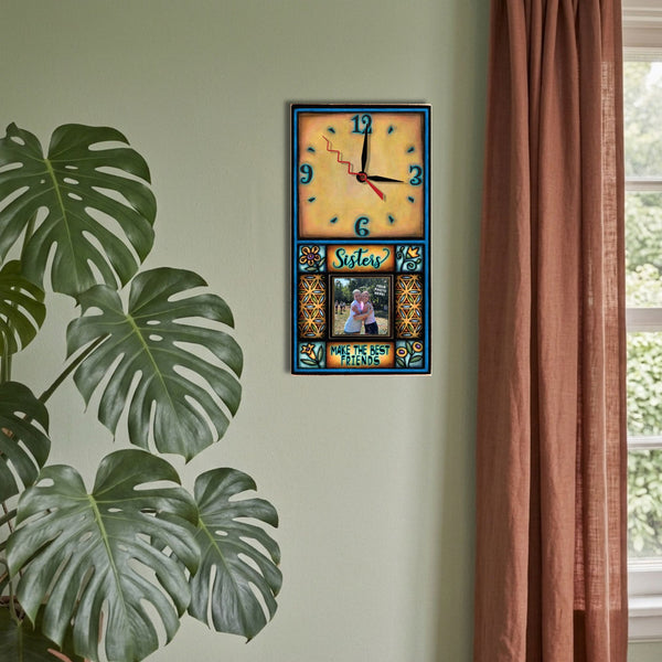Sisters Make the Best Friends picture frame wood wall clock hanging on a sage green wall beside monstera leaves and a rust linen curtain