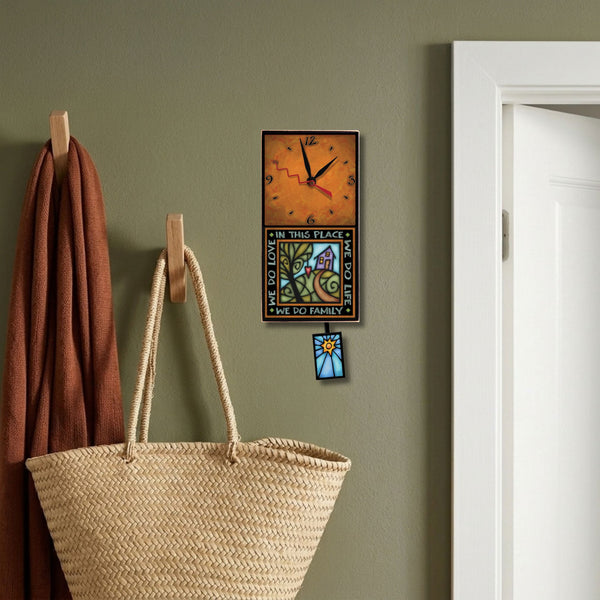 We Do Family handmade pendulum wall clock hanging on a muted olive green entryway wall beside wooden wall hooks