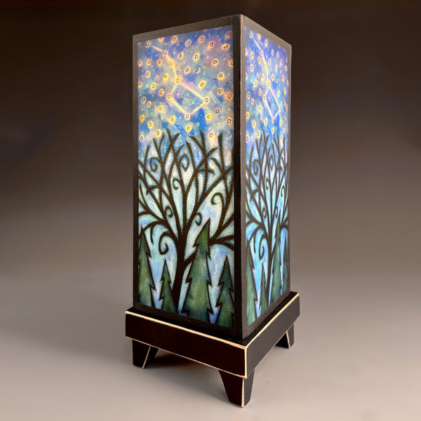 Constellations mood lamp with distressed wood base and glowing archival paper shade featuring swirling trees, evergreens, and a star-filled night sky.